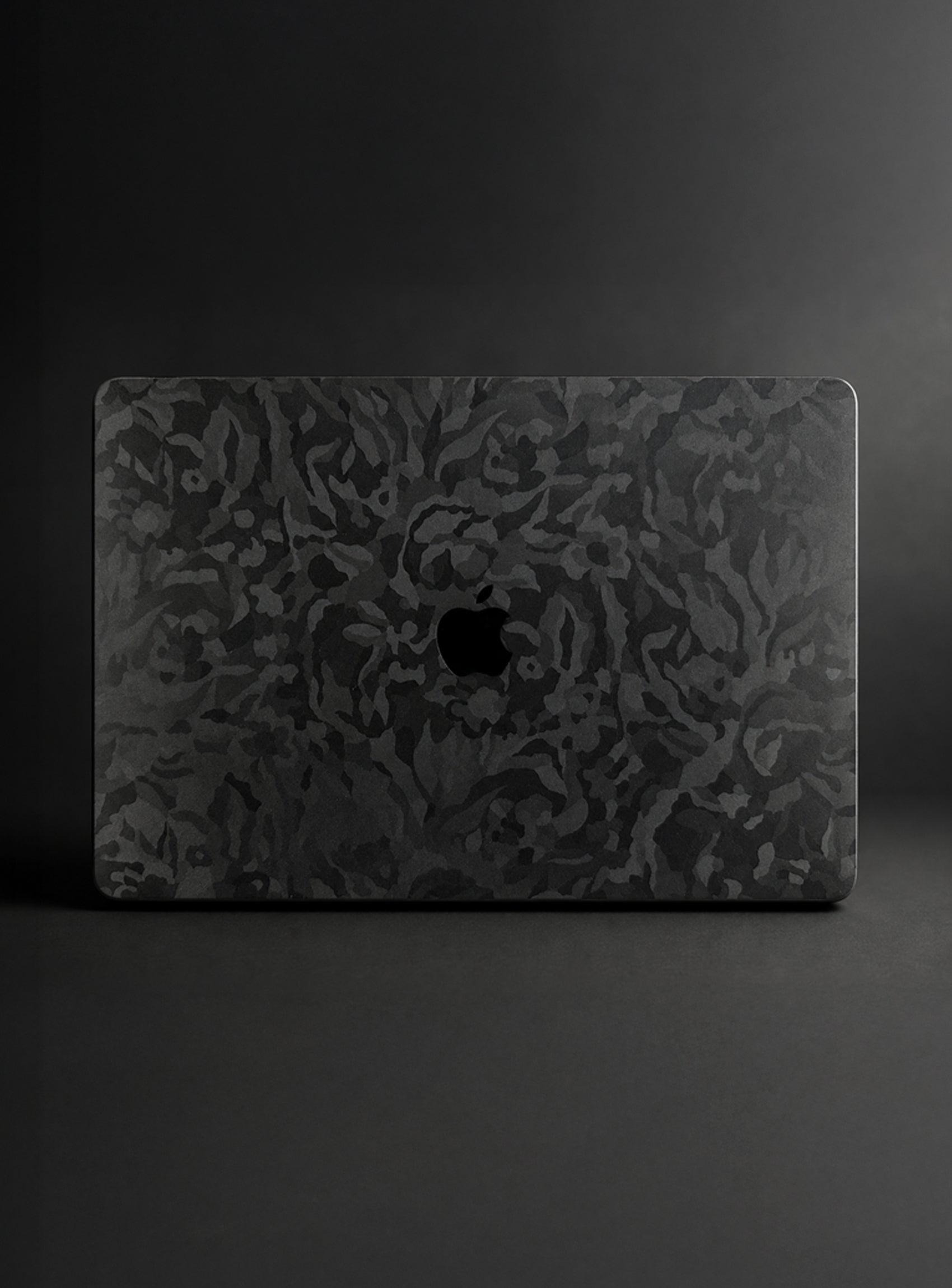 A camo textured apple macbook laptop skin or decal, featuring a black camo pattern and a cutout for the Apple logo, shown on a dark gray background.