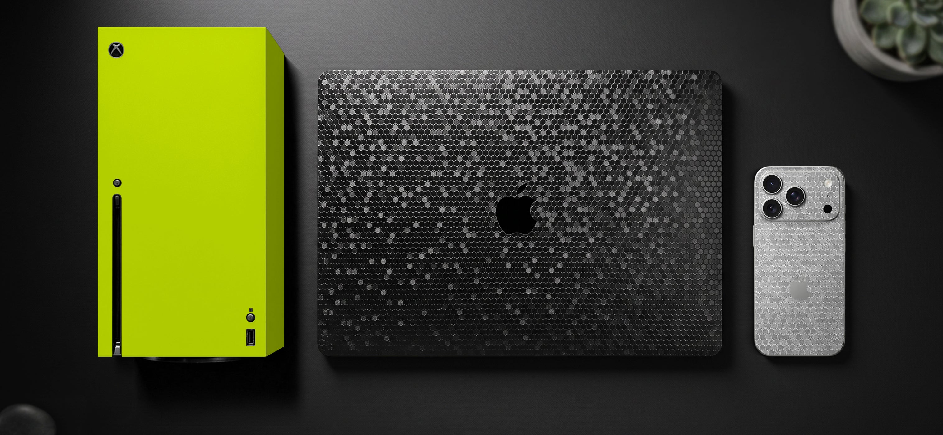 A flat-lay product presentation featuring a bright lime green Apple Vision Pro case on the left, a black laptop with a dark speckled skin in the center, and a silver smartphone with a light speckled skin on the right, all arranged on a dark surface.