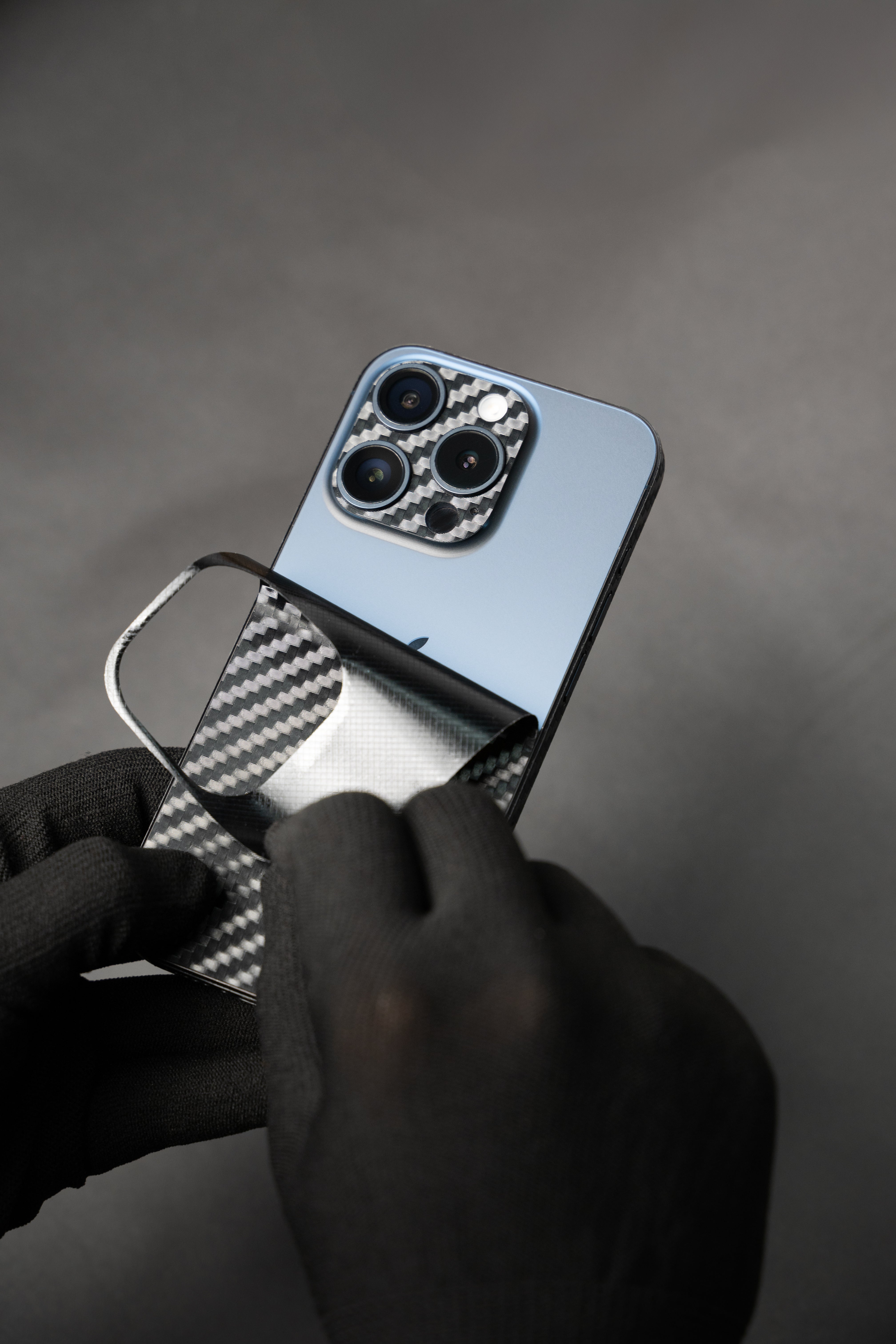 A close-up of a apple iphone 16 with a carbon fiber pattern skin being peeled off the back by gloved hands against a dark gray background.